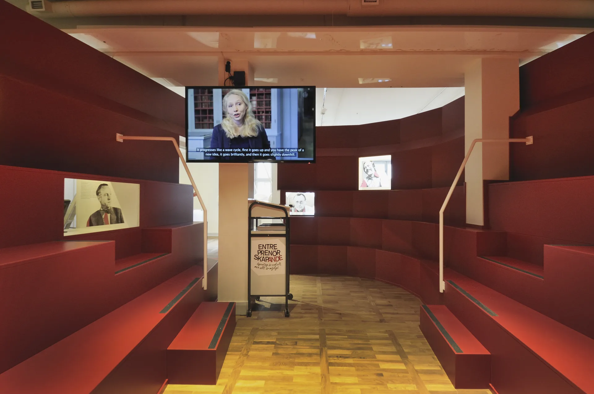 Utställningslokal med röda bänkar, ett tidningsställ och en tv som är påslagen med en intervju