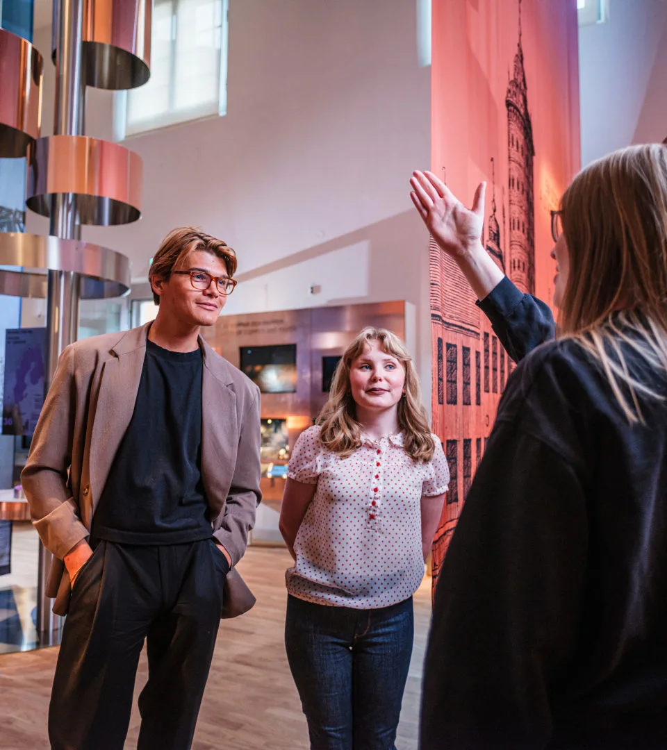 Pedagog pekar med handen mot ett föremål under en visning av utställningen PENGAR! för två personer som lyssnar.