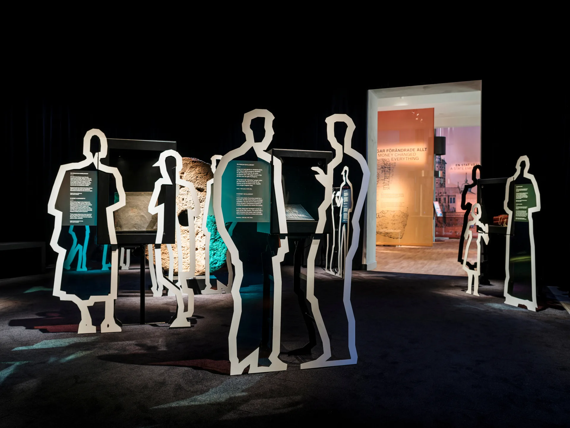 A view from the exhibition MONEY! featuring several black information panels mounted on life-sized white silhouettes of people. The silhouettes are cut out of cardboard or plastic and placed around display cases containing various objects, including a stone. In the background, an illuminated passage leads to another section of the exhibition, with the text “When Money Changed Everything” on the wall.