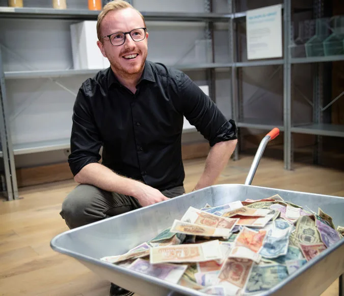 Pedagog sitter bakom en skottkärra med sedlar.