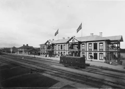 En svartvit bild på Ett stationshus bredvid några järnvägsspår. På husets tak finns två svenska flaggor.