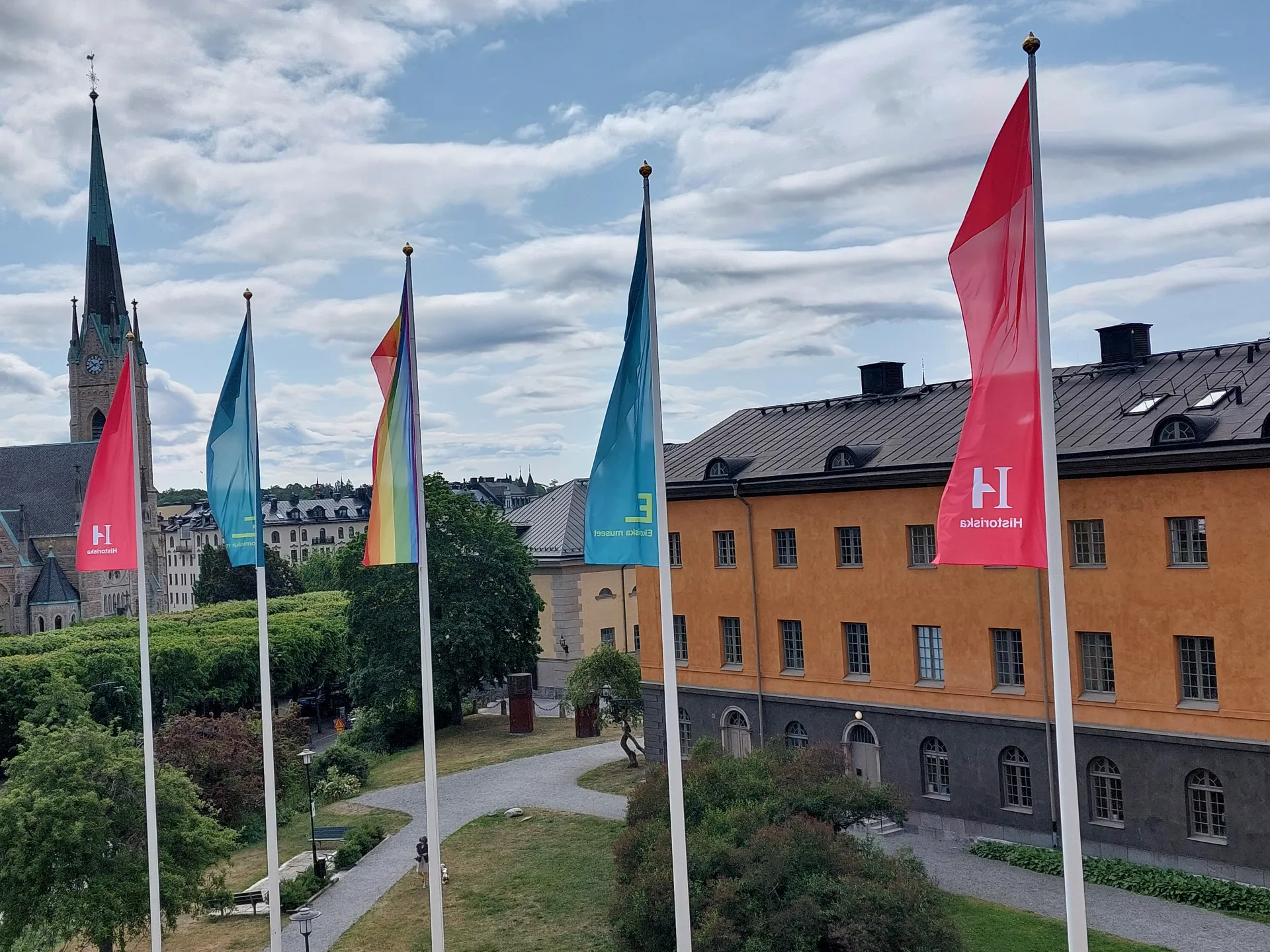 Prideflagga hissad bredvid flaggor för Ekonomiska museet och Historiska museet. Blå himmel.