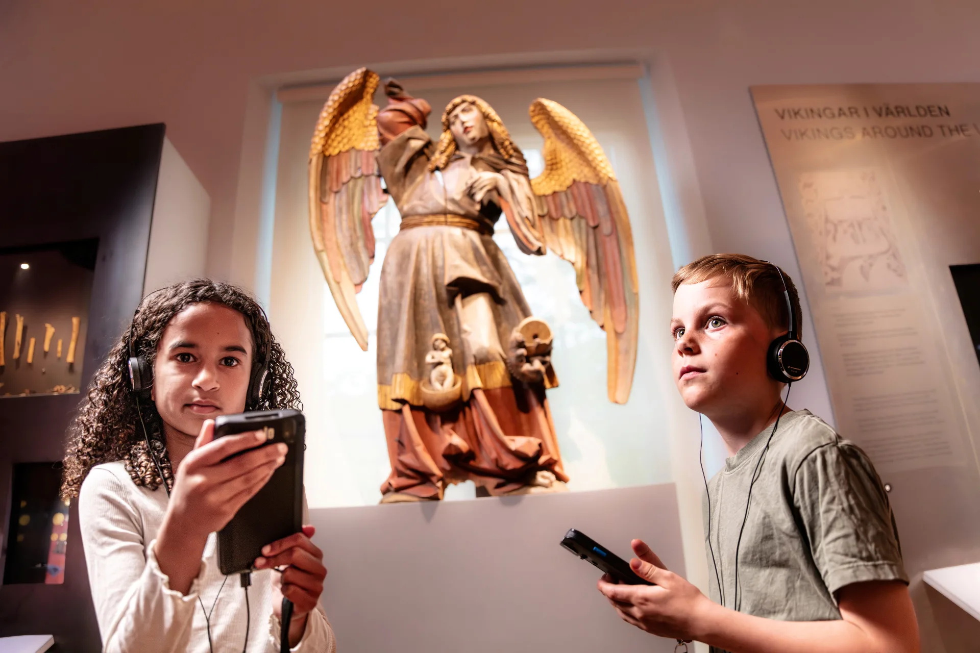 Två barn lyssnar på en audioguide vid en ängel i utställningen